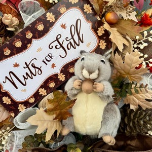 K&ouml;nnte beinhalten: Ein herbstliches Kranz mit einem Pl&uuml;sch-Eichh&ouml;rnchen, das eine Eichel h&auml;lt. Der Kranz enth&auml;lt ein Schild mit der Aufschrift "Nuts for Fall" mit Herbstbl&auml;ttern und Eichelmotiven. Die Farbpalette umfasst Braun-, Orange- und Gr&uuml;nt&ouml;ne mit dekorativen B&auml;ndern und Bl&auml;ttern.