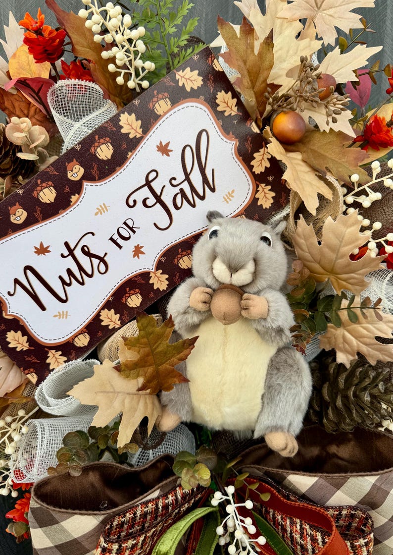 K&ouml;nnte beinhalten: Ein herbstliches Kranz mit einem Pl&uuml;sch-Eichh&ouml;rnchen, das eine Eichel h&auml;lt. Der Kranz enth&auml;lt ein Schild mit der Aufschrift "Nuts for Fall" mit Herbstbl&auml;ttern und Eichelmotiven. Die Farbpalette umfasst Braun-, Orange- und Gr&uuml;nt&ouml;ne mit dekorativen B&auml;ndern und Bl&auml;ttern.