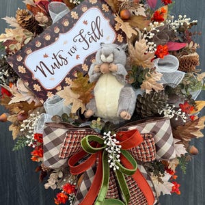 K&ouml;nnte beinhalten: Herbstliche Kranzdekoration mit einem Eichh&ouml;rnchen, das eine Nuss h&auml;lt, umgeben von Bl&auml;ttern, Tannenzapfen und einer Schleife. Auf dem Schild steht "Nuts for Fall".