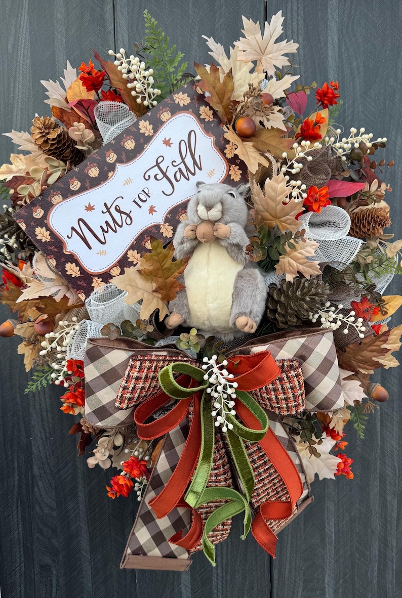 K&ouml;nnte beinhalten: Herbstliche Kranzdekoration mit einem Eichh&ouml;rnchen, das eine Nuss h&auml;lt, umgeben von Bl&auml;ttern, Tannenzapfen und einer Schleife. Auf dem Schild steht "Nuts for Fall".