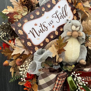 K&ouml;nnte beinhalten: Ein herbstliches Kranz mit einem Pl&uuml;sch-Eichh&ouml;rnchen, Herbstbl&auml;ttern und einem Schild mit der Aufschrift "Nuts for Fall". Der Kranz weist eine Vielzahl von Texturen und Farben auf, darunter Braun, Orange und Creme. Dekorative Eicheln und Tannenzapfen sind ebenfalls enthalten.