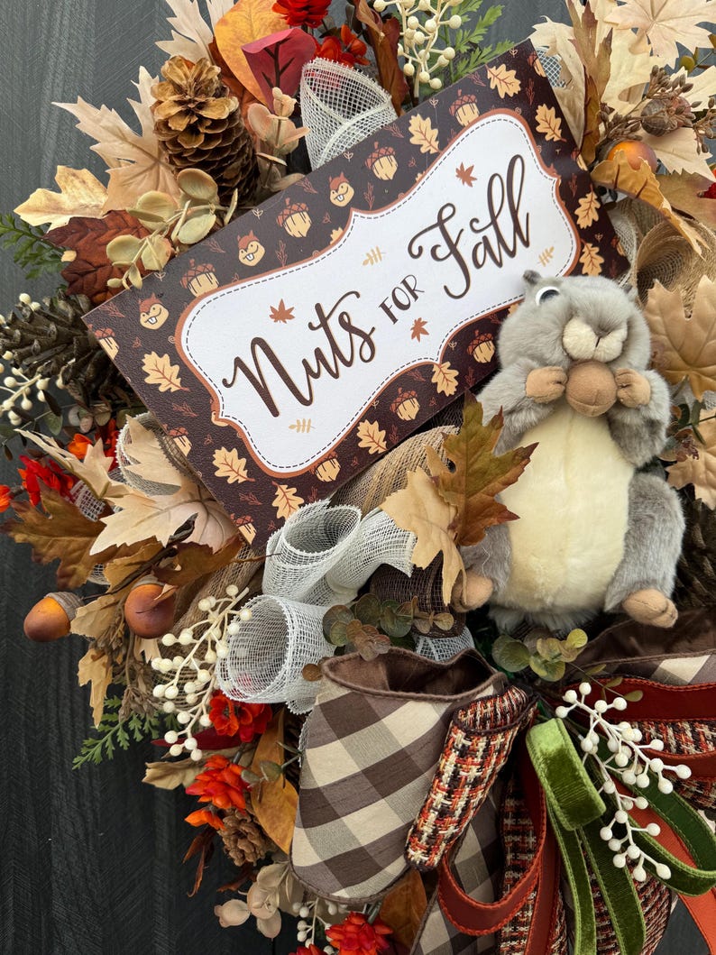 K&ouml;nnte beinhalten: Ein herbstliches Kranz mit einem Pl&uuml;sch-Eichh&ouml;rnchen, Herbstbl&auml;ttern und einem Schild mit der Aufschrift "Nuts for Fall". Der Kranz weist eine Vielzahl von Texturen und Farben auf, darunter Braun, Orange und Creme. Dekorative Eicheln und Tannenzapfen sind ebenfalls enthalten.