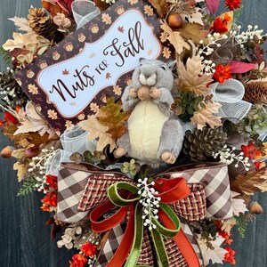 K&ouml;nnte beinhalten: Ein herbstliches Kranz mit einem Stofftier-Eichh&ouml;rnchen, das eine Nuss h&auml;lt. Der Kranz ist mit Herbstbl&auml;ttern, Tannenzapfen und einem Schild mit der Aufschrift "Nuts for Fall" geschm&uuml;ckt. Eine gro&szlig;e Schleife mit braunen, orangefarbenen und gr&uuml;nen B&auml;ndern sorgt f&uuml;r einen dekorativen Akzent.