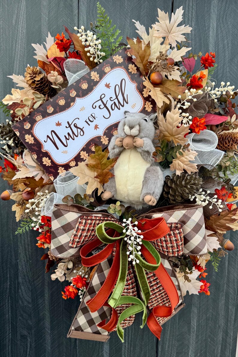 K&ouml;nnte beinhalten: Ein herbstliches Kranz mit einem Stofftier-Eichh&ouml;rnchen, das eine Nuss h&auml;lt. Der Kranz ist mit Herbstbl&auml;ttern, Tannenzapfen und einem Schild mit der Aufschrift "Nuts for Fall" geschm&uuml;ckt. Eine gro&szlig;e Schleife mit braunen, orangefarbenen und gr&uuml;nen B&auml;ndern sorgt f&uuml;r einen dekorativen Akzent.