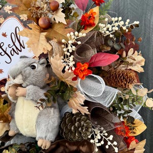 K&ouml;nnte beinhalten: Ein herbstliches Kranz mit einem grauen Pl&uuml;sch-Eichh&ouml;rnchen, das eine Eichel h&auml;lt. Der Kranz enth&auml;lt Herbstbl&auml;tter, orangefarbene Blumen, Tannenzapfen und ein Schild mit der Aufschrift "Fall". Braune und wei&szlig;e B&auml;nder und Akzente vervollst&auml;ndigen das Design.