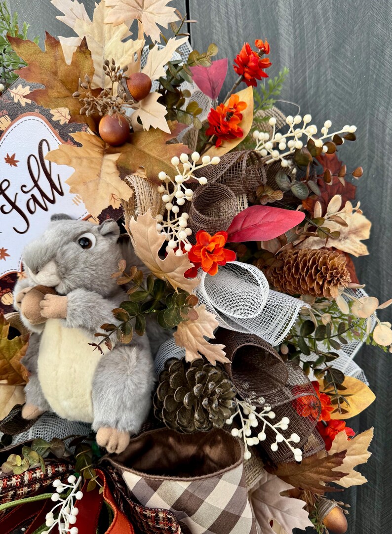 K&ouml;nnte beinhalten: Ein herbstliches Kranz mit einem grauen Pl&uuml;sch-Eichh&ouml;rnchen, das eine Eichel h&auml;lt. Der Kranz enth&auml;lt Herbstbl&auml;tter, orangefarbene Blumen, Tannenzapfen und ein Schild mit der Aufschrift "Fall". Braune und wei&szlig;e B&auml;nder und Akzente vervollst&auml;ndigen das Design.