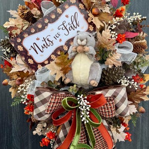 K&ouml;nnte beinhalten: Ein herbstliches Kranz mit einem Pl&uuml;sch-Eichh&ouml;rnchen, das eine Nuss h&auml;lt. Der Kranz zeigt Herbstbl&auml;tter, Tannenzapfen und ein Schild mit der Aufschrift "Nuts for Fall". Eine gro&szlig;e Schleife mit braunen, orangefarbenen und gr&uuml;nen B&auml;ndern rundet das Design ab.