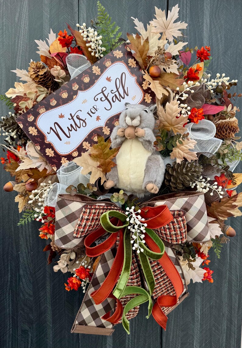 K&ouml;nnte beinhalten: Ein herbstliches Kranz mit einem Pl&uuml;sch-Eichh&ouml;rnchen, das eine Nuss h&auml;lt. Der Kranz zeigt Herbstbl&auml;tter, Tannenzapfen und ein Schild mit der Aufschrift "Nuts for Fall". Eine gro&szlig;e Schleife mit braunen, orangefarbenen und gr&uuml;nen B&auml;ndern rundet das Design ab.