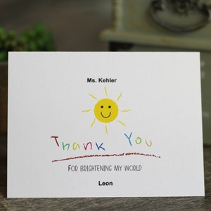 以下が含まれることがあります： 黄色い笑顔の太陽のイラストと、カラフルな手書き文字で「Thank You」と書かれた白いグリーティングカード。「FOR BRIGHTENING MY WORLD」というテキストと「Leon」という名前も含まれています。