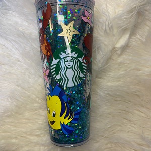 May include: A clear, double-walled tumbler with a green straw, featuring a Starbucks logo and a design of characters from The Little Mermaid. The tumbler is filled with teal glitter and decorative elements, including a starfish and floral accents.