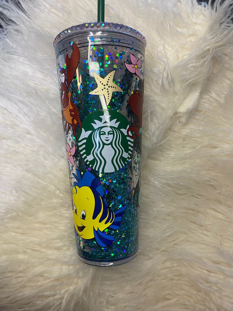 May include: A clear, double-walled tumbler with a green straw, featuring a Starbucks logo and a design of characters from The Little Mermaid. The tumbler is filled with teal glitter and decorative elements, including a starfish and floral accents.