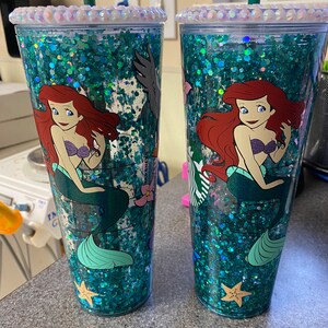 May include: Two clear tumblers with a glittery teal interior and a mermaid design. The tumblers feature a cartoon image of Ariel, the Little Mermaid, and are adorned with iridescent rhinestones around the rim. Each tumbler has a green straw.