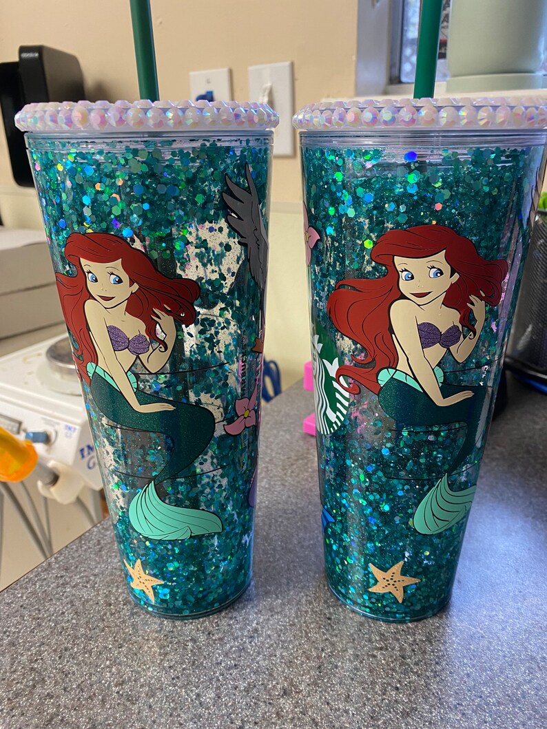 May include: Two clear tumblers with a glittery teal interior and a mermaid design. The tumblers feature a cartoon image of Ariel, the Little Mermaid, and are adorned with iridescent rhinestones around the rim. Each tumbler has a green straw.