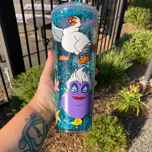May include: A clear, double-walled tumbler with a turquoise glitter interior and a green straw. The tumbler features cartoon images of a white seagull and a purple Ursula the sea witch. The rim is decorated with clear rhinestones.