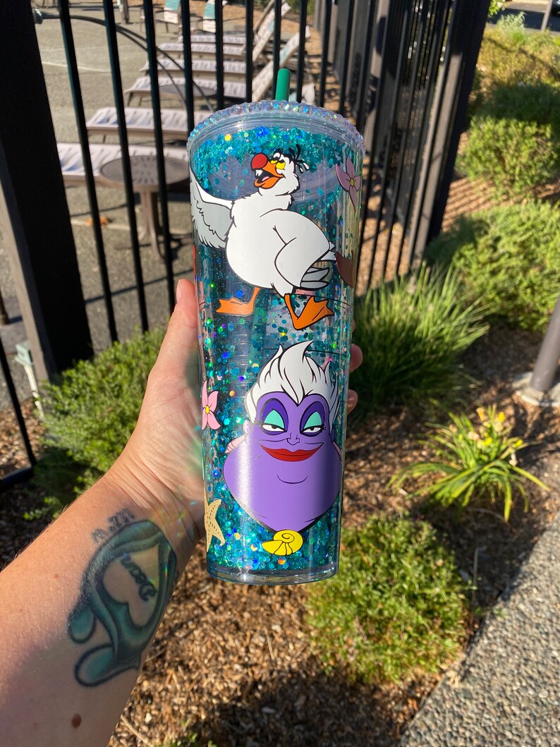 May include: A clear, double-walled tumbler with a turquoise glitter interior and a green straw. The tumbler features cartoon images of a white seagull and a purple Ursula the sea witch. The rim is decorated with clear rhinestones.
