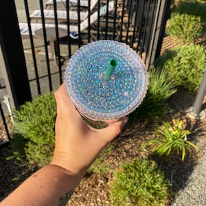 May include: A clear, iridescent tumbler with a bedazzled lid and a green straw. The cup is held in a hand, with a background of green bushes and a black fence.