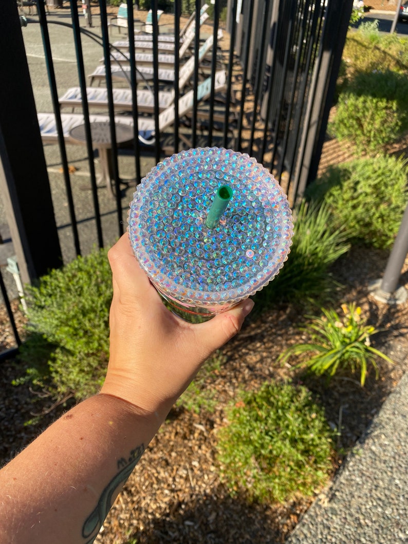 May include: A clear, iridescent tumbler with a bedazzled lid and a green straw. The cup is held in a hand, with a background of green bushes and a black fence.