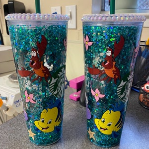 May include: Two clear tumblers with teal glitter and characters from The Little Mermaid. Each tumbler features Sebastian the crab and Flounder the fish, with pink flowers and starfish. The lids are decorated with iridescent rhinestones and have a green straw.