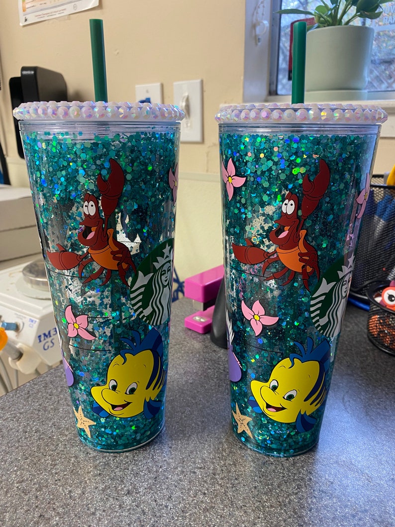 May include: Two clear tumblers with teal glitter and characters from The Little Mermaid. Each tumbler features Sebastian the crab and Flounder the fish, with pink flowers and starfish. The lids are decorated with iridescent rhinestones and have a green straw.