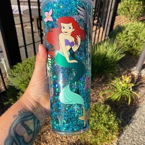 May include: A clear, double-walled tumbler filled with blue glitter and featuring a cartoon image of Ariel from The Little Mermaid. The tumbler has a green straw and is decorated with pink flowers and a starfish.