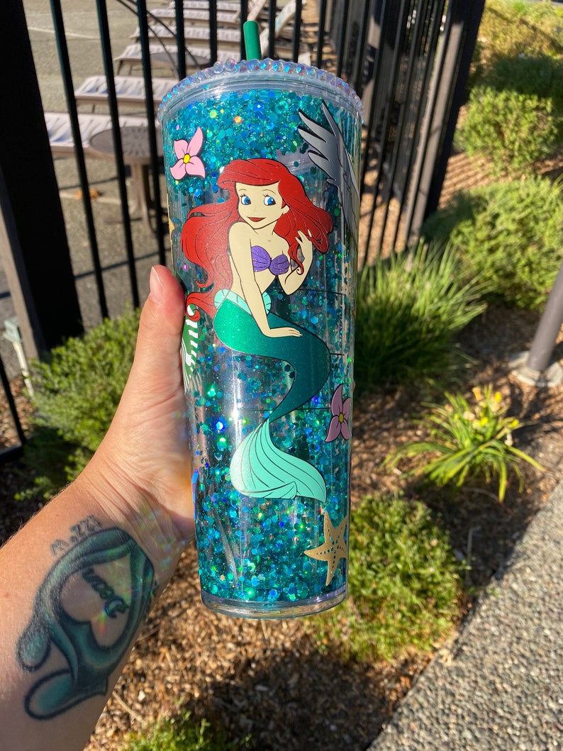 May include: A clear, double-walled tumbler filled with blue glitter and featuring a cartoon image of Ariel from The Little Mermaid. The tumbler has a green straw and is decorated with pink flowers and a starfish.