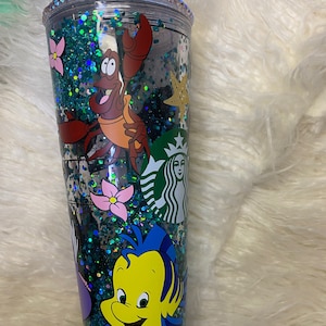 May include: A clear Starbucks tumbler with a green straw, filled with teal glitter and character decals. The tumbler features illustrations of Sebastian the crab, Flounder, and other characters from The Little Mermaid. The rim is decorated with rhinestones.