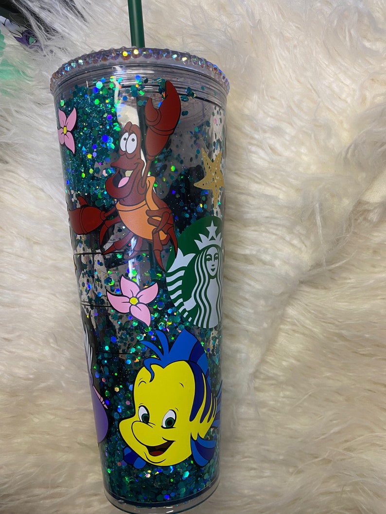 May include: A clear Starbucks tumbler with a green straw, filled with teal glitter and character decals. The tumbler features illustrations of Sebastian the crab, Flounder, and other characters from The Little Mermaid. The rim is decorated with rhinestones.
