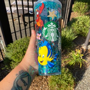 May include: A clear, double-walled tumbler filled with blue glitter and featuring cartoon characters from The Little Mermaid, including Sebastian and Flounder. A Starbucks logo is also visible. The tumbler has a green straw and lid.