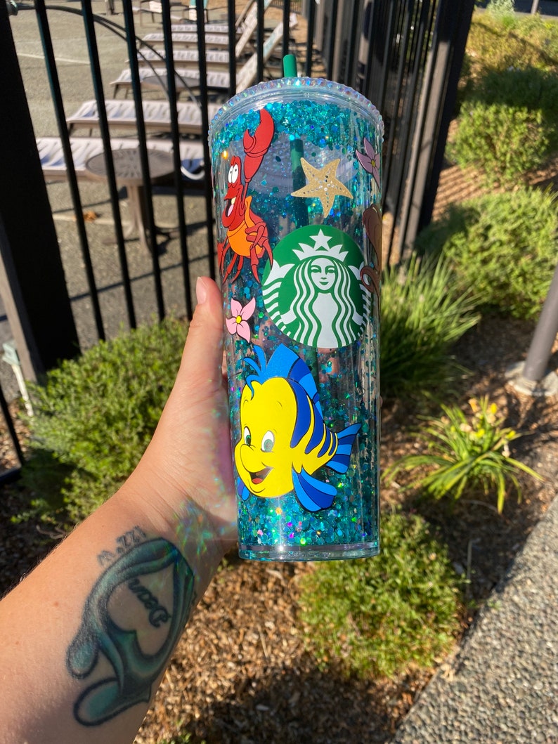 May include: A clear, double-walled tumbler filled with blue glitter and featuring cartoon characters from The Little Mermaid, including Sebastian and Flounder. A Starbucks logo is also visible. The tumbler has a green straw and lid.