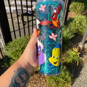 May include: A clear, double-walled tumbler with a green straw, filled with blue glitter and featuring cartoon characters from The Little Mermaid. The characters include Sebastian, Flounder, and Ursula, with pink flower accents.