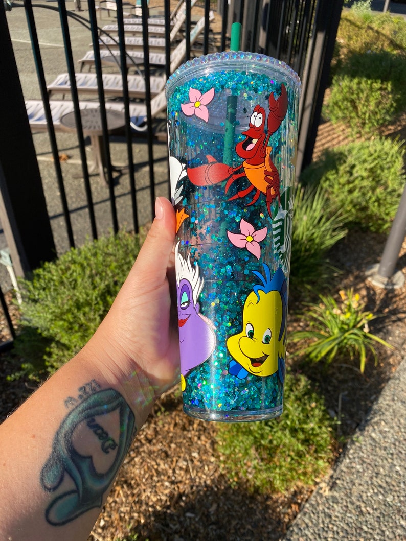 May include: A clear, double-walled tumbler with a green straw, filled with blue glitter and featuring cartoon characters from The Little Mermaid. The characters include Sebastian, Flounder, and Ursula, with pink flower accents.