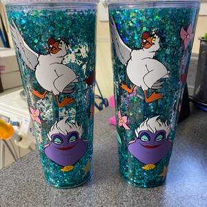 May include: Two clear tumblers with teal glitter and character designs. Each tumbler features a white seagull, a purple Ursula, and pink flowers. The rims are decorated with clear rhinestones, and each has a green straw.