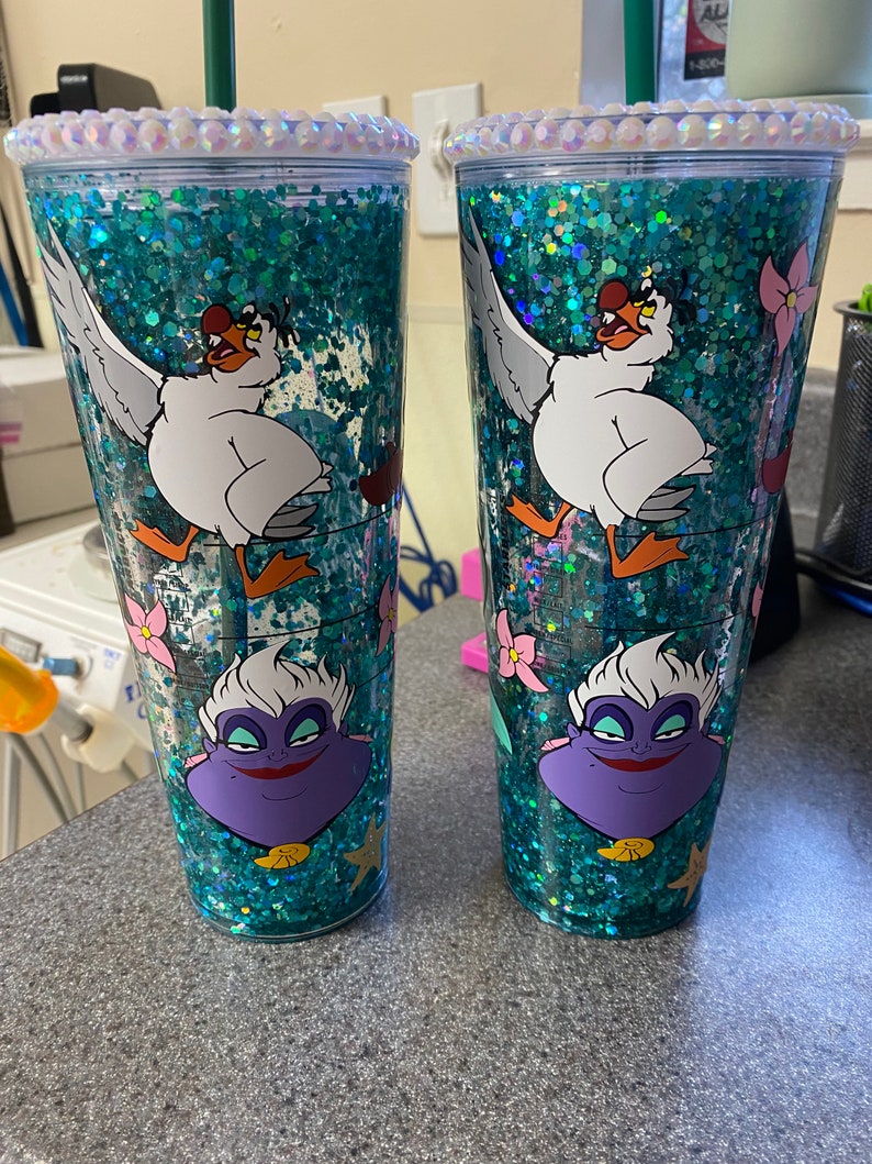 May include: Two clear tumblers with teal glitter and character designs. Each tumbler features a white seagull, a purple Ursula, and pink flowers. The rims are decorated with clear rhinestones, and each has a green straw.