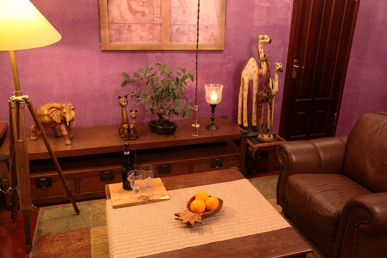 May include: A wooden elephant figurine, two wooden cat figurines, and two wooden giraffe figurines are displayed on a wooden shelf. A wooden table with a bowl of oranges sits in front of the shelf. A brown leather armchair is in the background.
