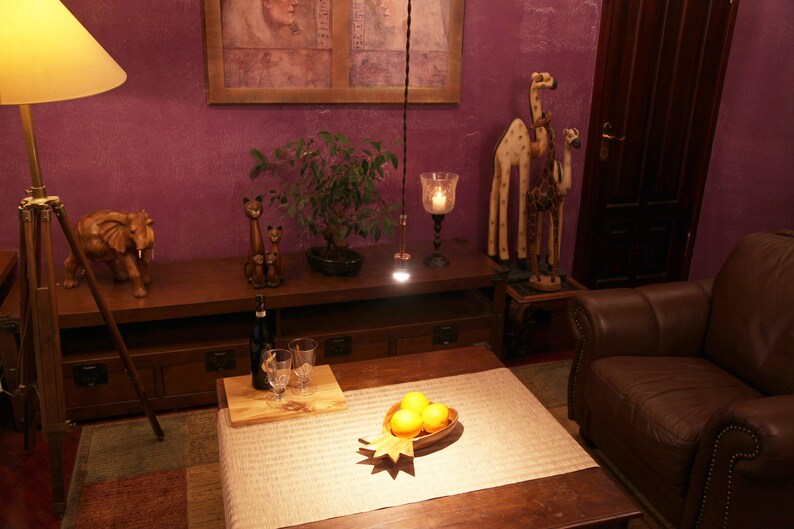May include: A living room interior with a wooden table, a brown leather armchair, and a wooden tripod lamp with a lampshade. There are wooden animal figurines on the table, including an elephant and giraffes. A potted bonsai tree is on the table. There are three oranges in a wooden bowl on a table with a white tablecloth.