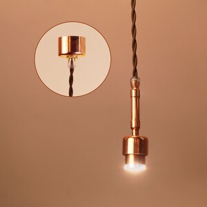 May include: A copper pendant light fixture with a brown braided cord. The light is on and emitting a bright white light.
