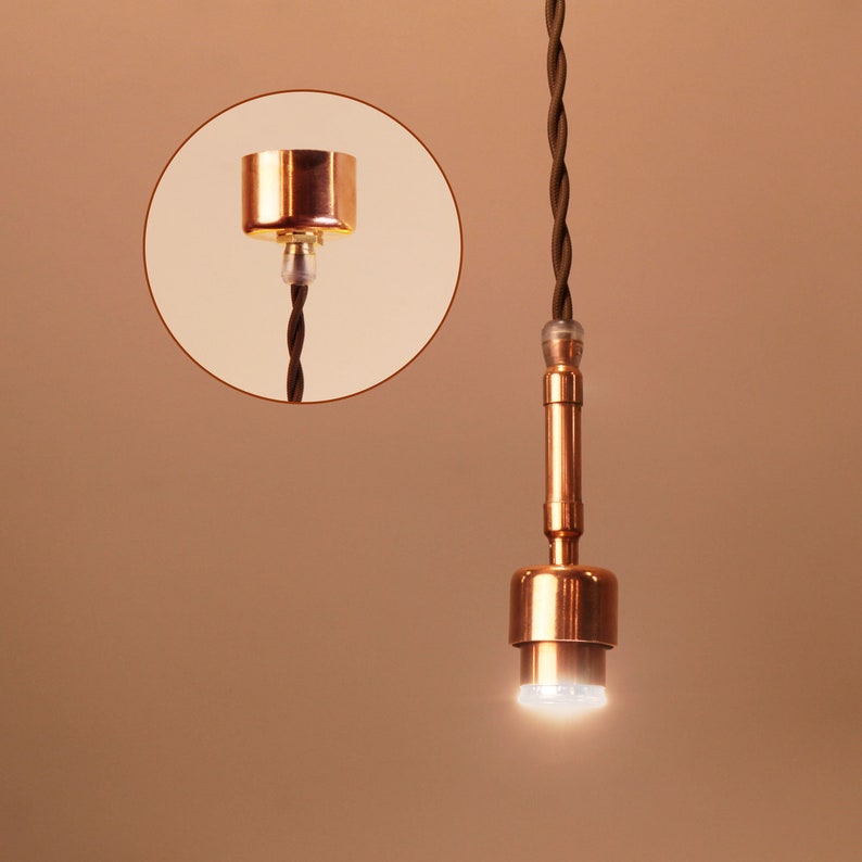 May include: A copper pendant light fixture with a brown braided cord. The light is on and emitting a bright white light.
