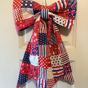 May include: A patriotic, quilted bow in red, white, and blue. The bow features various patterns, including polka dots, stars, stripes, and zigzags. It is designed to hang and is likely a decorative item for a door or wall.