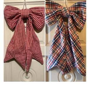 May include: Two fabric bows hanging vertically. The left bow features a red and white gingham check pattern. The right bow displays a plaid pattern in red, white, and blue with small floral accents. Both bows have two long, pointed tails and a central knot.