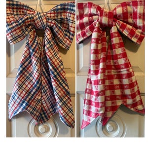 May include: Two fabric bows with long tails. The left bow features a blue, red, and white plaid pattern. The right bow features a red and white gingham check pattern with small repeating fruit motifs printed within the white squares.