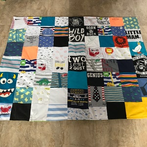 Pode incluir: Uma colcha de retalhos feita de roupas de bebê com vários desenhos e frases coloridos, incluindo "Wild Boy", "Two Legit 2 Quit", "Cool Like Daddy" e "Little Man".