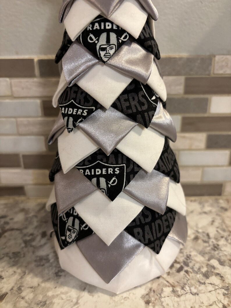May include: A white, silver, and black fabric Christmas tree with the Raiders logo and the word "Raiders" repeated throughout.