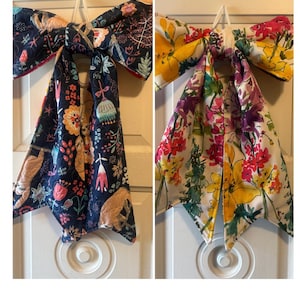 May include: Two fabric bows hanging vertically. The left bow features a dark navy background with colorful illustrations of sloths, flowers, and foliage. The right bow has a white background with a vibrant, multi-colored floral print pattern.