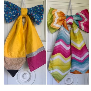 May include: Two large fabric bows with hanging tails. The left bow features a blue school-themed print with a yellow pencil design, including a tan tip, silver band, and pink eraser. The right bow has a colorful chevron pattern in pink, yellow, and blue.