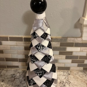 May include: A black and white fabric Christmas tree with a black ornament on top. The tree is decorated with the Las Vegas Raiders logo.