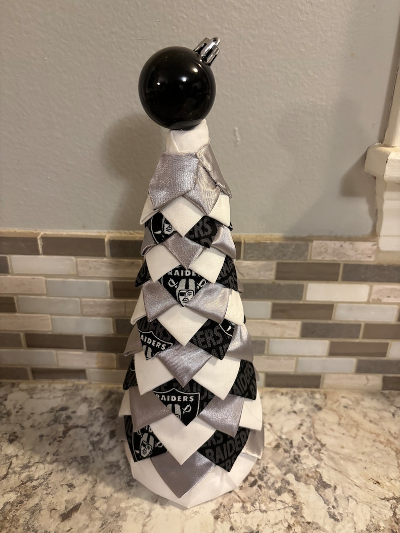May include: A black and white fabric Christmas tree with a black ornament on top. The tree is decorated with the Las Vegas Raiders logo.