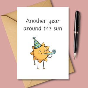 Puede incluir: Una tarjeta de felicitación blanca con el texto "Another year around the sun" y un sol de dibujos animados con un gorro de fiesta y soplando una bocina. La tarjeta está junto a un sobre marrón y un bolígrafo negro.