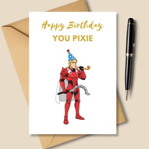 May include: A white greeting card with the text "Happy Birthday YOU PIXIE" in gold lettering. The card features a cartoon illustration of a person in red armor and a party hat. A black pen and a brown envelope are also in the image.