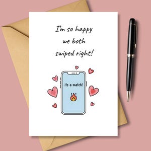 May include: A white greeting card with the text "I'm so happy we both swiped right!" features a smartphone graphic with the words "its a match!" and a flame icon. Pink hearts surround the phone. A black pen and a brown envelope are also in the image.