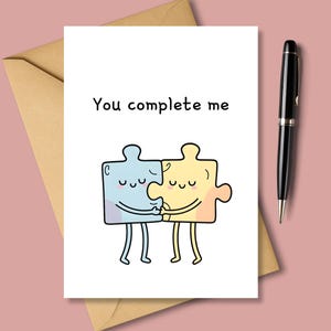 May include: A greeting card with the text "You complete me" above two cartoon puzzle pieces hugging. The puzzle pieces are blue and yellow with closed eyes. A black pen and a brown envelope are also in the image.